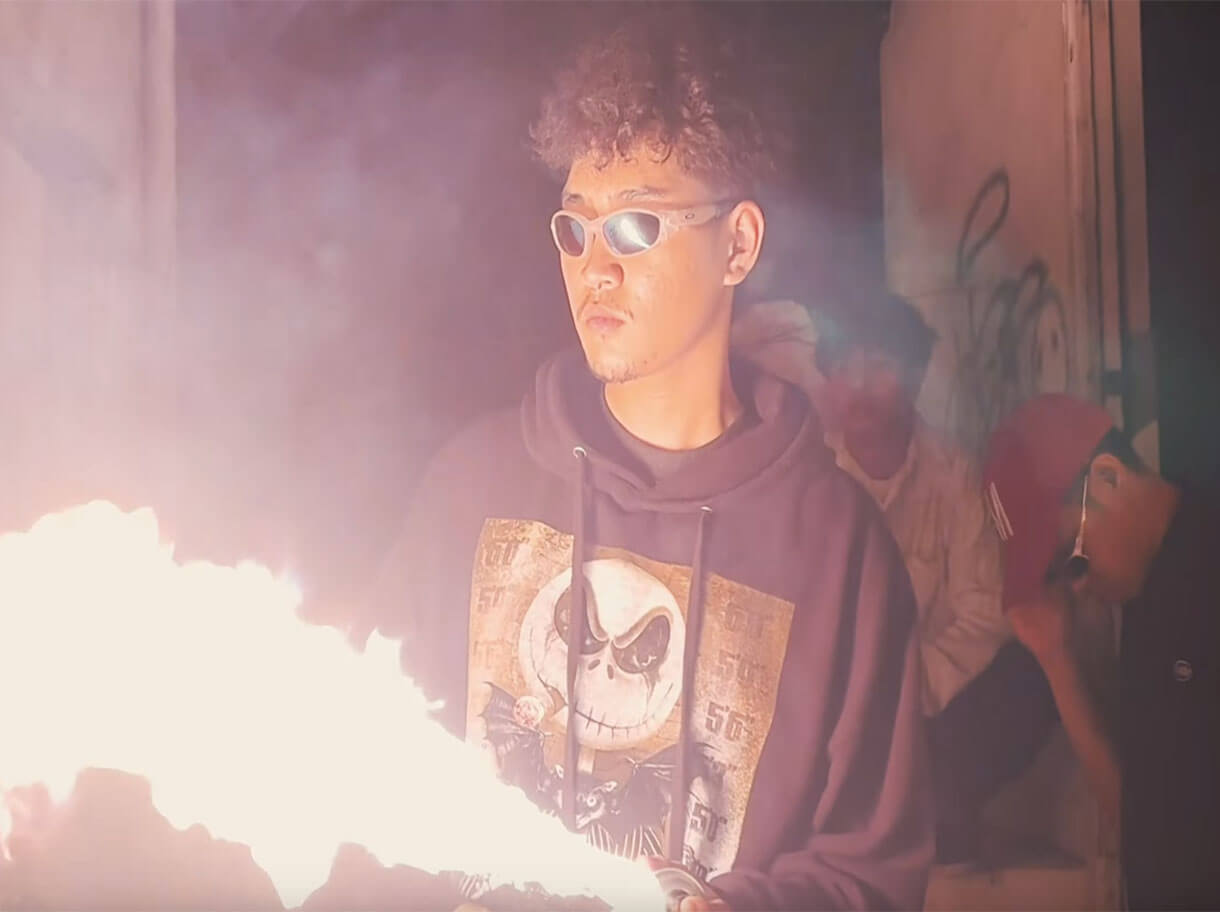 Person in a hoodie and sunglasses holding a spray can and lighter that emits a large flame, with bystanders in the background as intro of THA KREW music video.