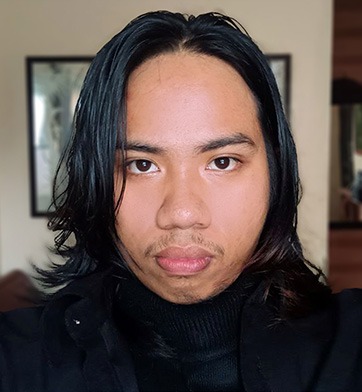 Close-up portrait of a person with shoulder-length dark hair, neutral expression, wearing dark clothing in an indoor setting.